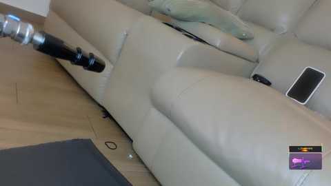A video of a modern living room with a beige leather sectional sofa, a wooden floor, a black remote control, and a silver electrical cord extending from the left.