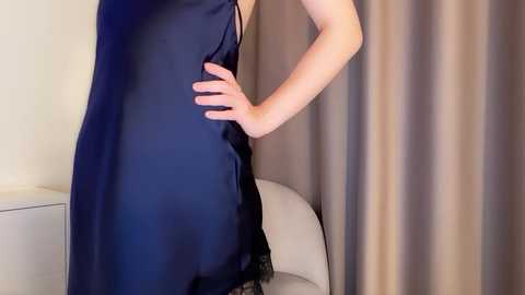 Media: Video of a woman in a dark blue, sleeveless satin dress with a thin black lace trim, standing in front of beige curtains and a white chair, left hand on her hip.