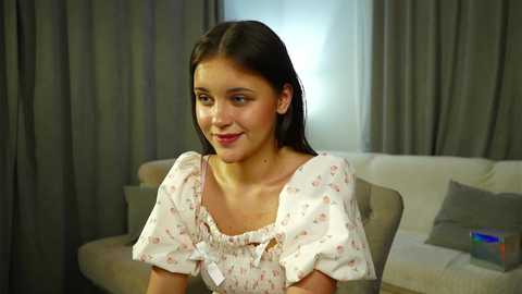 Video of a young woman with straight, dark hair, wearing a white floral dress, seated on a beige sofa in a dimly lit living room with gray curtains and a gray pillow.