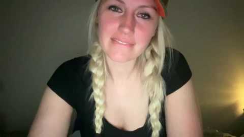 Video of a young Caucasian woman with fair skin, long blonde braids, and a black t-shirt. She smiles slightly with a neutral expression. Background is dimly lit, with a yellow lamp on the right.