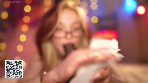 Media: A video of a blonde woman with glasses, wearing a dark hat, eating a white cake with red icing. The background is blurred with colorful lights, suggesting a festive setting.
