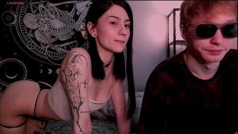Video of a woman with black hair and tattoos, wearing a white crop top, and a man with glasses and a black shirt, both on a bed, in a dimly lit room.