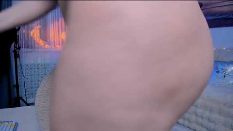 A video of a nude person's rounded buttocks, captured from a side angle, showing smooth skin and a subtle glow. The background features a dimly lit, modern room with a textured wall and a cozy, patterned rug.