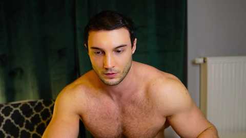 Video of a shirtless, muscular, light-skinned man with short dark hair, gazing downward, in a dimly lit room with a dark curtain and white radiator.