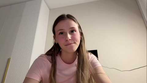 Video of a young Caucasian girl with long, straight brown hair, wearing a light pink t-shirt, standing in a plain, beige-walled room with a vertical, textured white panel on the left.