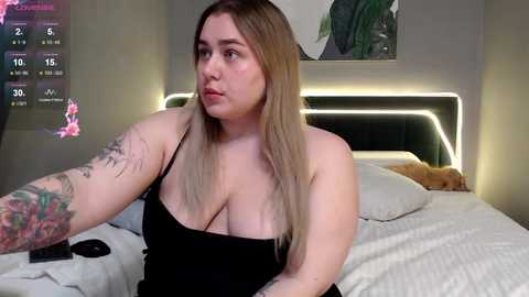 Video of a plus-sized woman with long blonde hair, large breasts, and a sleeve tattoo, wearing a black dress, sitting on a bed with white sheets in a modern, dimly lit bedroom.