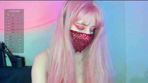 Video of a pale-skinned woman with long, straight, pink hair, wearing a red glitter mask and red eyeshadow, set against a pastel-colored wall.