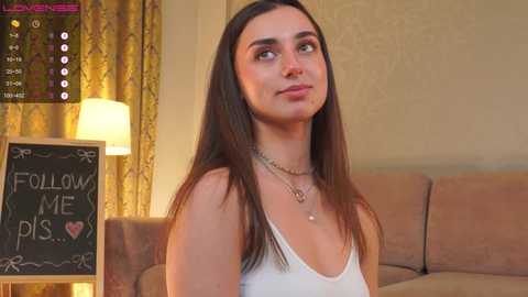 Video of a young woman with long brown hair, wearing a white tank top and necklace, standing next to a chalkboard sign saying \"FOLLOW ME\" in a cozy, beige-lit room with a lamp and curtains.