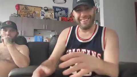 Video of two shirtless men in a living room, one in a black basketball jersey, smiling, the other in a baseball cap. Background features a fridge, cabinets, and sports memorabilia.