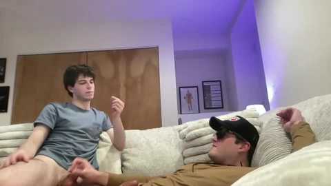 Video of two young men: one in a blue shirt, smoking, the other in a baseball cap, lounging on a beige couch, in a dimly lit living room.