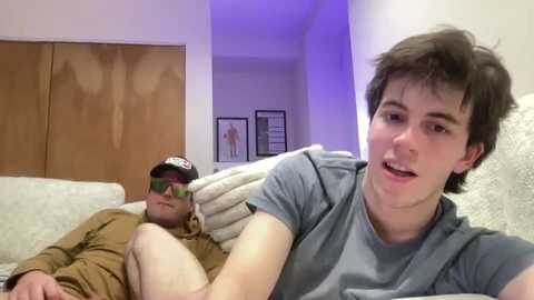 Video of two young men in a bedroom: one lounging on a bed in brown clothing, and the other, wearing a gray t-shirt, smiling.