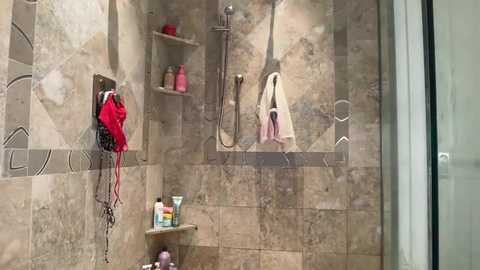Media: A modern, beige-tiled shower with a glass door, featuring a metallic handheld showerhead, red towel, and a pink bathrobe hanging, and toiletries on a shelf.