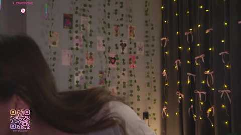 Video of a dimly lit bedroom with a person's back, long brown hair, and a bed with white sheets. Walls adorned with fairy lights and small green plants.