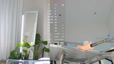 Video of a modern, minimalist room with a large, transparent, elevated bed, a potted plant, a vertical LED light panel, and a white curtain on the left.