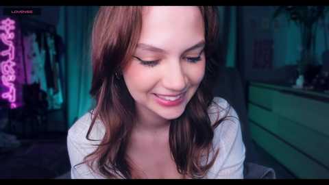 Video of a smiling, light-skinned woman with long, wavy auburn hair, wearing a light grey top, in a dimly lit room with neon lights and a green dresser in the background.