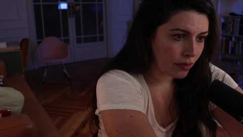 A video of a young woman with long black hair, fair skin, and a contemplative expression, sitting in a dimly lit room with wooden furniture and a bookshelf in the background.