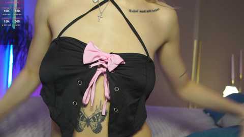 Video of a woman wearing a black corset with a pink bow, revealing a butterfly tattoo on her abdomen. Background shows a dimly lit room with a bed and a blurred TV screen displaying \"VOD\" text.
