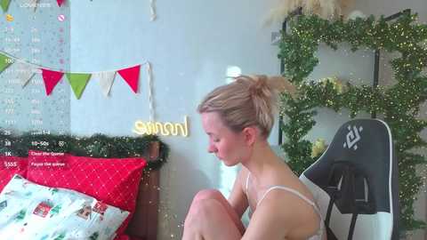 Video of a blonde woman with fair skin, wearing a white bra, sitting on a gaming chair in a festive room with Christmas decorations, including a calendar, garland, and a \"swamp\" sign.