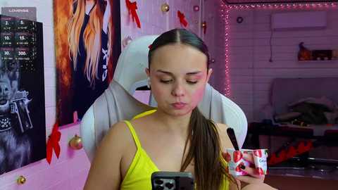 Video of a young woman with light skin and straight, long brown hair in a ponytail, wearing a yellow tank top, taking a selfie in a brightly decorated bedroom.