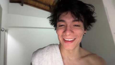 Video of a young Asian man with short, tousled black hair, wearing a white towel, smiling widely, with a white door and wooden ceiling in the background.
