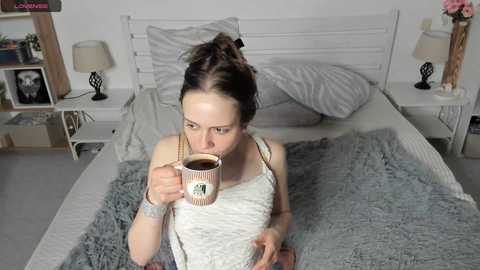 Video of a young woman with light skin and brown hair in a messy bun, wearing a striped tank top, sipping coffee from a mug, in a cozy, white-themed bedroom with a gray blanket, pillows, and nightstands.