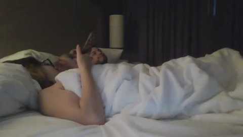 A video of a couple in bed, the woman lying on her side, partially covered by white sheets, watching TV with a remote in her hand. The man is asleep beside her, covered in sheets.