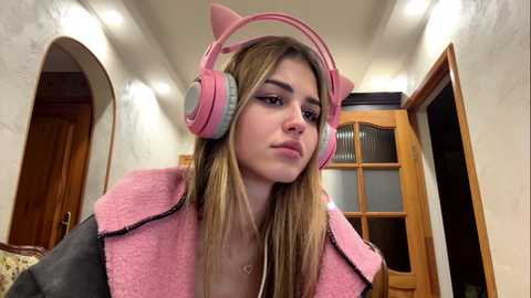 A young woman with long blonde hair, wearing pink headphones and a pink fluffy jacket, stands indoors with wooden doors and archways in the background.