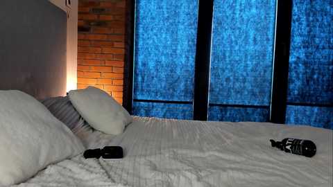 Video of a modern bedroom with a white bed, black bottles, brick wall, and large dark-framed windows with blue-tinted curtains, creating a cozy yet minimalist ambiance.