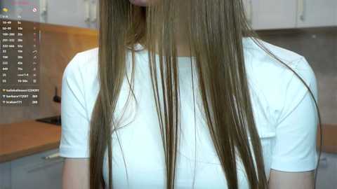Video of a young woman with long, straight, light brown hair, wearing a white T-shirt, standing in a modern kitchen with wooden cabinets and stainless steel appliances.