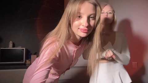Video of two young girls with long blonde hair in a dimly lit room. One wears a pink shirt, the other a white dress.