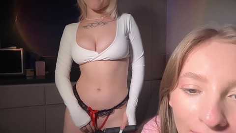 Video of a blonde woman in a white crop top and black lace panties, standing in a dimly lit room. Another woman with blonde hair and pink shirt partially visible.