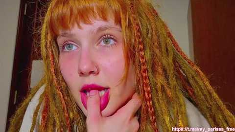 Media: Video of a pale-skinned woman with bright orange dreadlocks, wearing pink lipstick, and holding her finger to her mouth, set against a beige background.