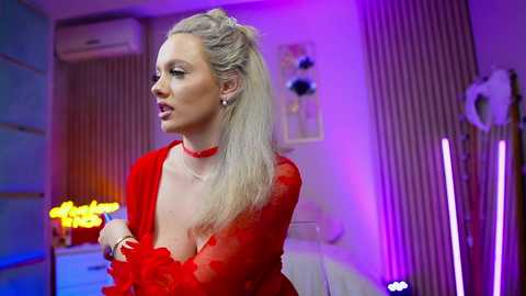 Video of a blonde woman in a red, sheer, low-cut dress with a choker, standing in a dimly lit room with vertical striped wallpaper.