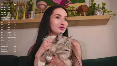 Video of a young woman with long black hair, holding a gray and white cat, seated on a green sofa, surrounded by mushroom decorations and a colorful painting in a cozy room.