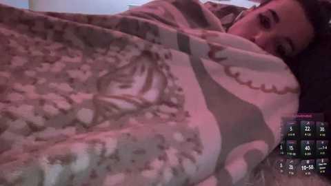 Video of a young woman with dark hair, lying under a patterned blanket, partially visible on a bed, with a TV screen showing a weather forecast in the background.