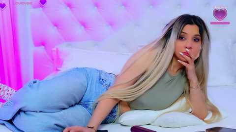 Video of a light-skinned woman with long, blonde hair, wearing a green tank top and light blue jeans, lying on her stomach on a bed with a white headboard and pink lighting.