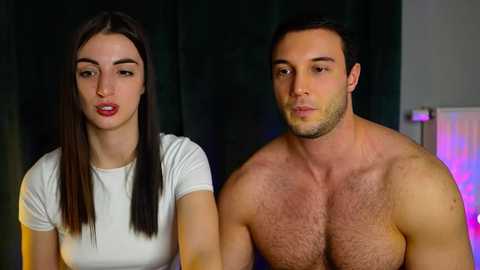 Video of a young, fair-skinned woman with long brown hair, wearing a white top, standing next to a shirtless, muscular man with short dark hair, against a dark curtain backdrop.