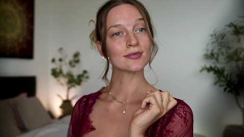 Video of a fair-skinned woman with auburn hair in a red lace top, standing indoors, with a blurred background featuring plants and a bed.
