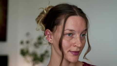 A video of a fair-skinned woman with blonde hair in a bun, wearing a burgundy top, smiling softly, against a blurred, beige background.