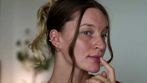 Video of a fair-skinned, freckled woman with auburn hair tied back, wearing a gold hoop earring and gold ring, with a thoughtful expression, against a blurred green background.