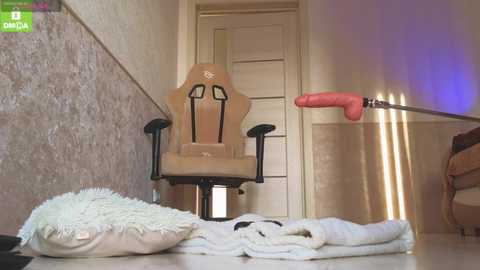 Video of a beige gaming chair with black armrests, a white plush pillow, and a white towel on a tiled floor. In the background, a pink dildo rests on a stand, next to a closed door.