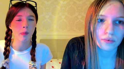 Media: Video of two young women with fair skin, one with braided hair and glasses, the other with straight hair, both wearing casual clothes, in a softly lit room with beige wallpaper.