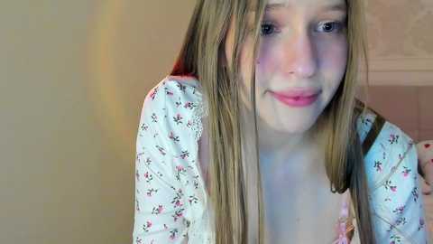 Media: Video of a young Caucasian girl with long blonde hair, fair skin, and a soft smile, wearing a floral-patterned shirt, indoors with a blurred beige wall background.