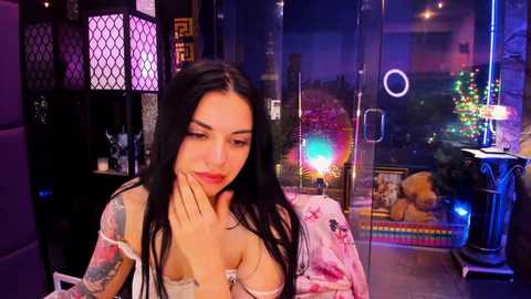 Video of a young woman with long black hair, fair skin, and red lipstick, wearing a floral robe, seated indoors with colorful neon lights and a stuffed animal in the background.