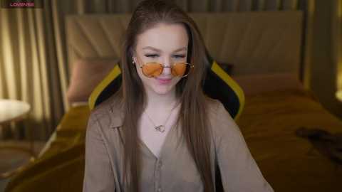 Video of a smiling, fair-skinned woman with long, straight brown hair, wearing sunglasses, beige shirt, and sitting on a bed with a gold and black patterned headboard.