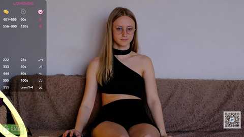 Media: A video of a young Caucasian woman with long blonde hair, wearing a black one-shoulder crop top and shorts, sitting on a brown couch against a plain wall.