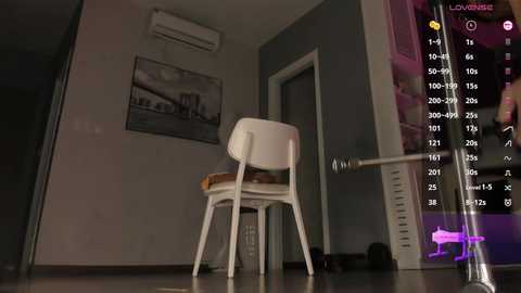 Video of a minimalist, dimly-lit room with a white chair, wall art, and a digital screen displaying a list of names and numbers.