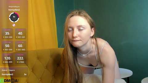 A video of a young, fair-skinned woman with long, straight brown hair, wearing a sheer white top, leaning on a bed, against a teal and yellow wall.