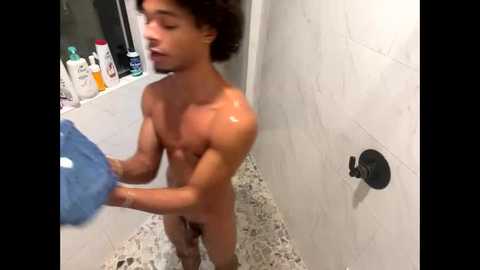 Video of a young, muscular, nude man with curly hair, soaping up in a white-tiled shower with a speckled floor.