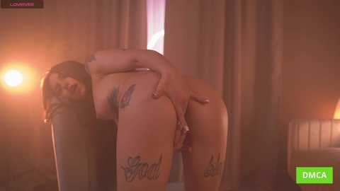 Media: Video of a nude woman with light skin, tattoos on her thighs, bending over, revealing her shaved vulva, in a dimly lit room with curtains and a lit candle.